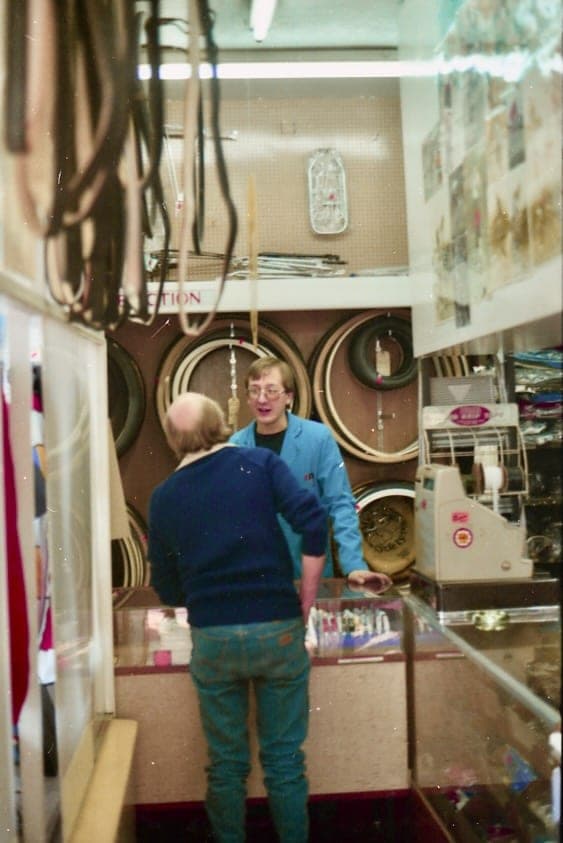 Two people at the back of the shop under a wall of tubular tyres hung in loops, one facing the camera and one turned away towards the glass counter.