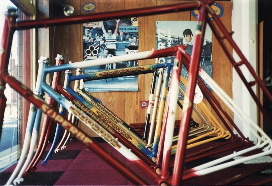 Half a dozen unbuilt frames in different colours leaning together, with Colnago posters pinned on the panelled wall behind them.