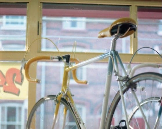 A pale gold racing bicycle standing in the shop window photographed from inside, the brick terrace across the road showing through the glass behind it.