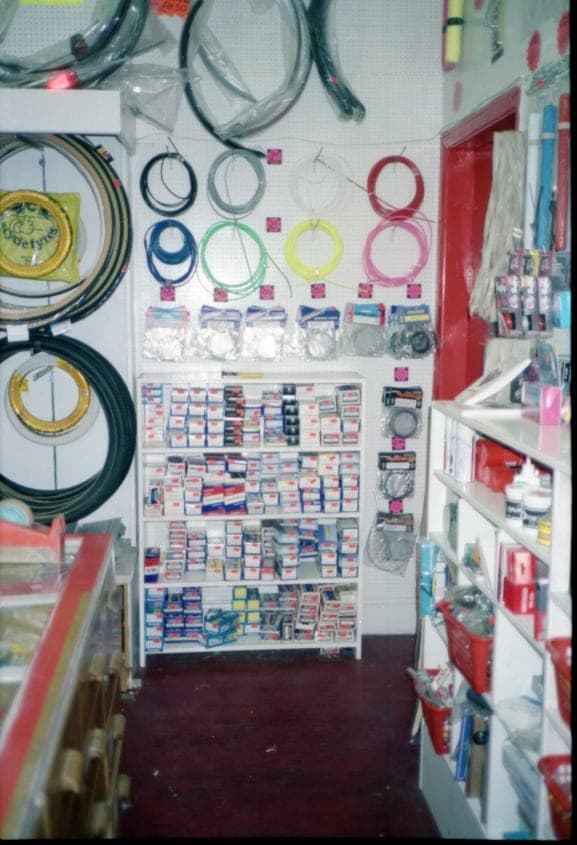 A shop wall hung with coiled brake and gear cables in packets above a rack of boxed inner tubes and small parts.