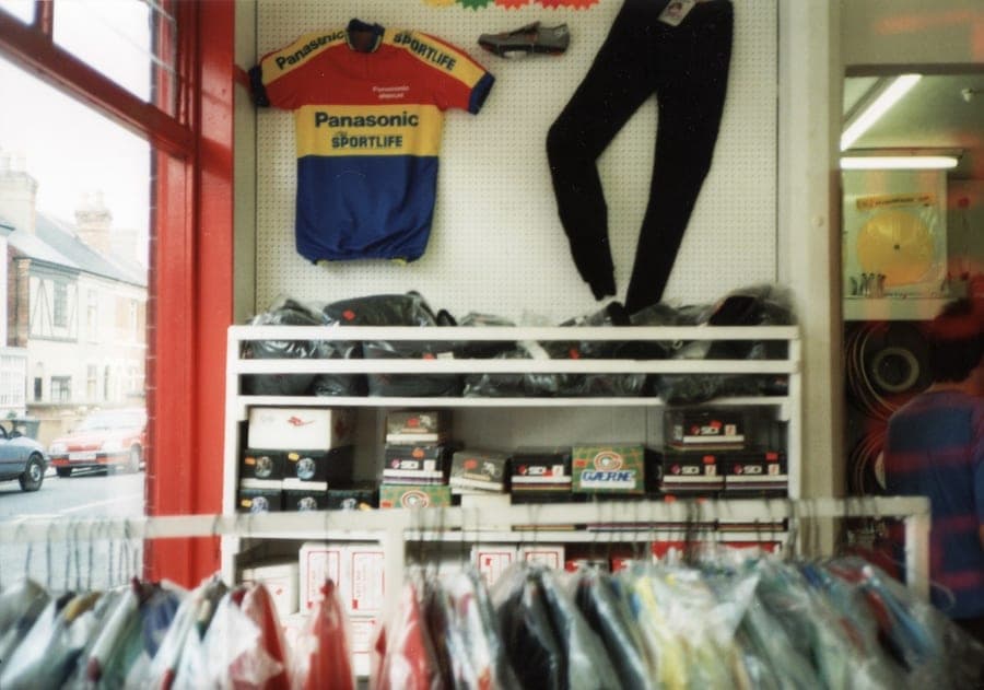 Racing jerseys and shorts on a pegboard above a rail of packaged clothing, with the shop window and the street beyond to the left.