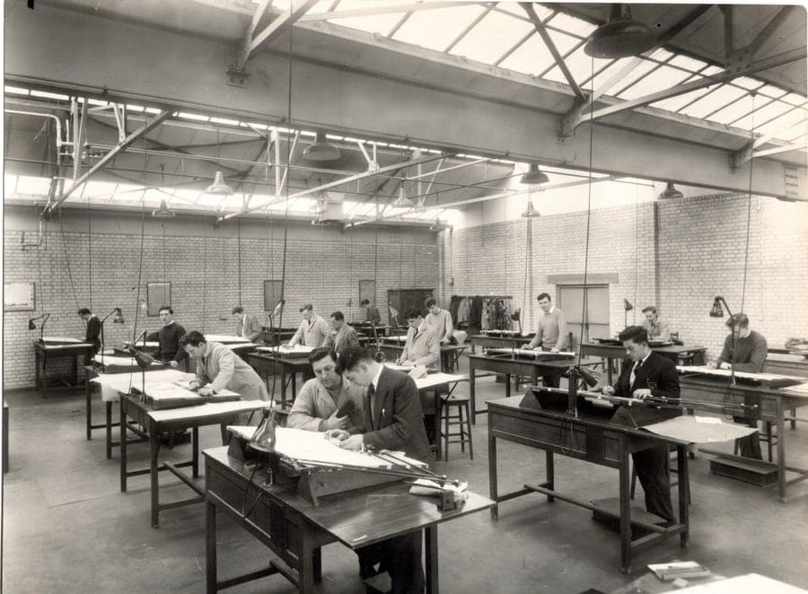 Rows of drafting tables in a top-lit industrial drawing office, a dozen men working at them, in black and white.