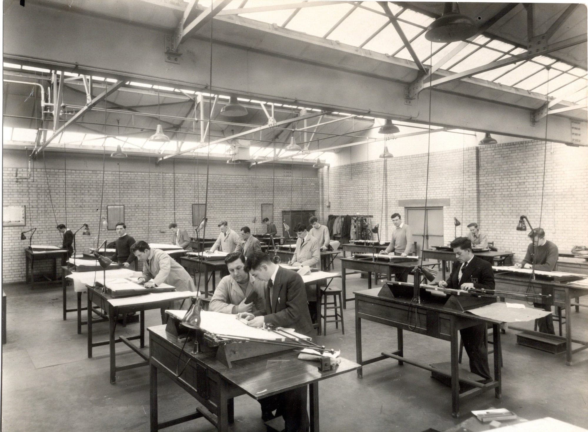 Rows of drafting tables in a top-lit industrial drawing office, a dozen men working at them, in black and white.