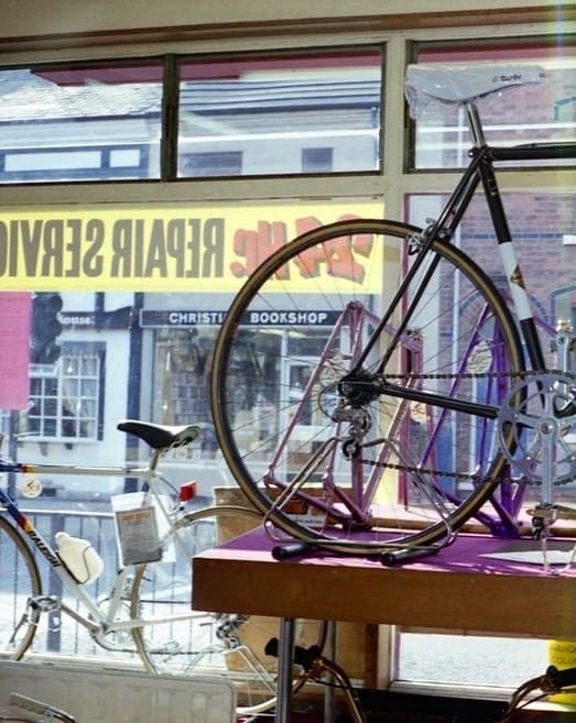 A bare frame and forks resting on a bench in the shop window, with 'Cycle Repair Service' lettered in reverse across the glass and a Christian bookshop visible across the road.