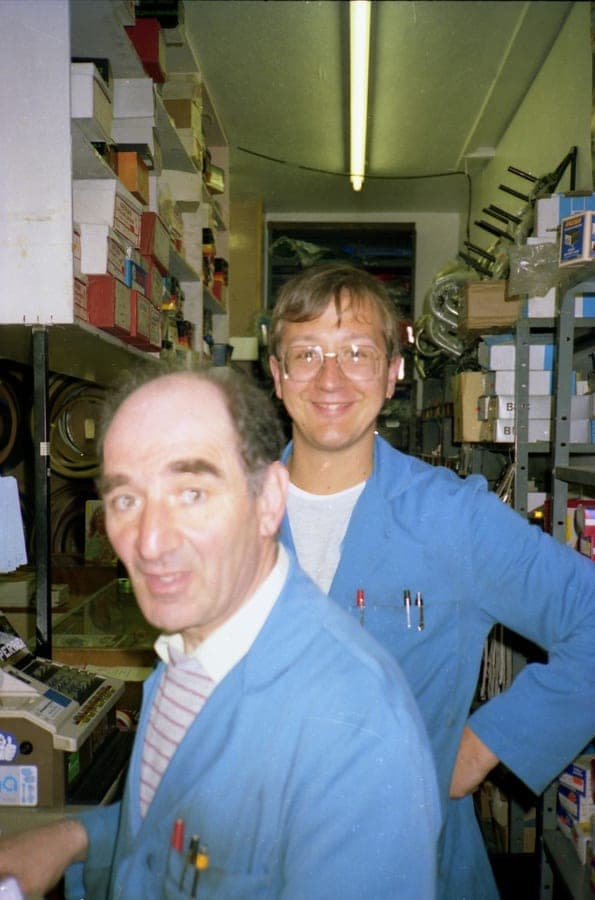 Sid in a shirt and tie beside Kevin in a blue workshop coat, at the counter among boxes of stock.