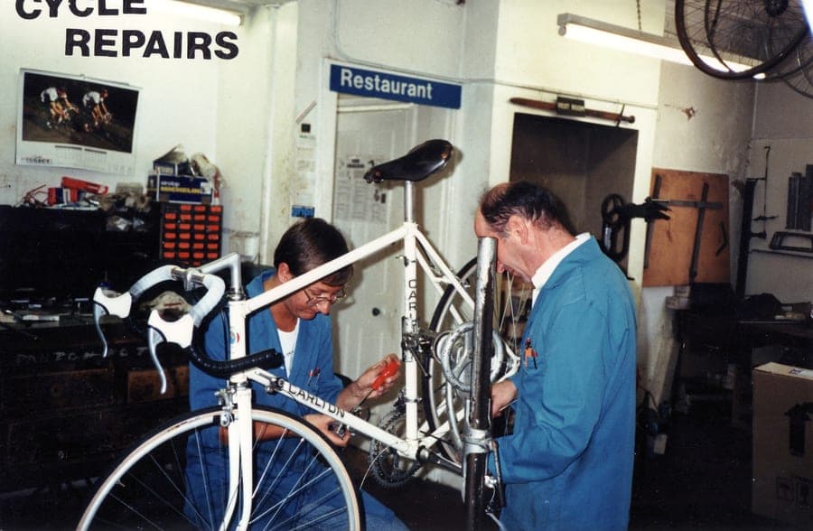 Two mechanics working on a white Carlton road bike under a sign reading 'CYCLE REPAIRS'.