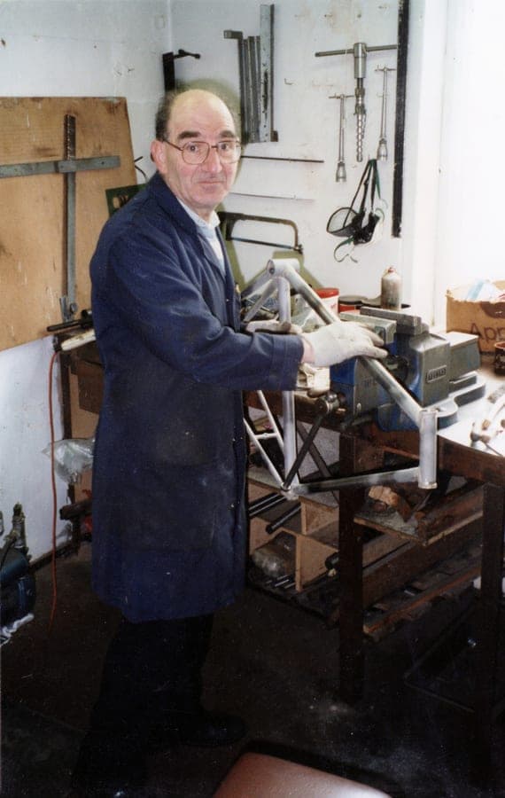 A frame builder in a navy workshop coat stands at a jig with a bare frame clamped into it, turning to face the camera.