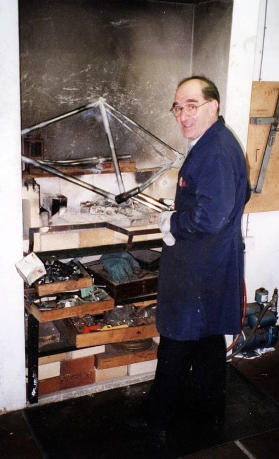 A frame held over a brick brazing hearth, the wall behind it black with soot, the builder looking round from the bench.