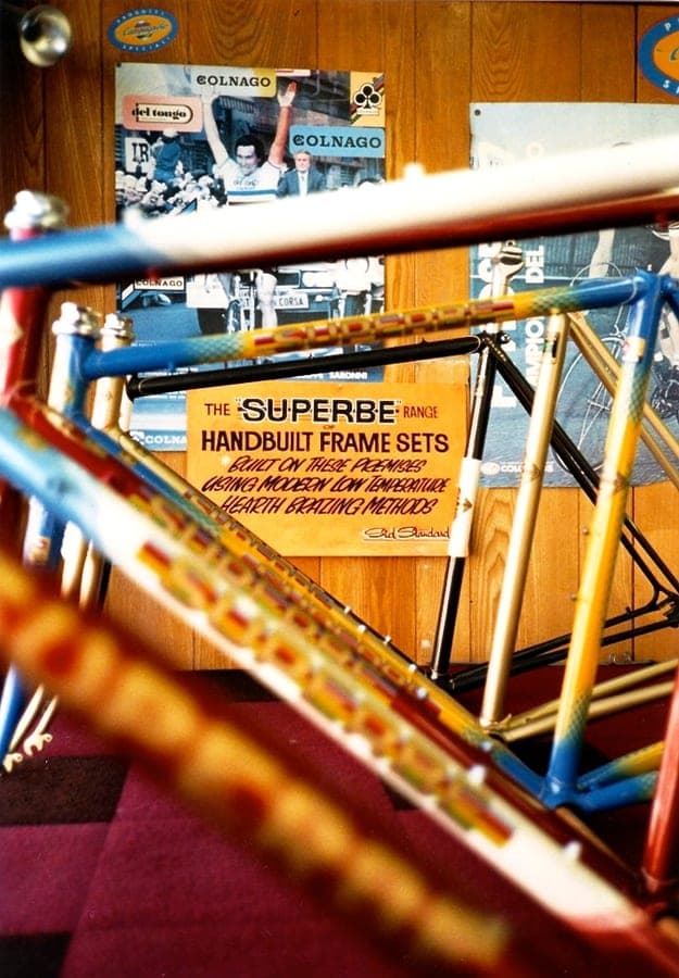 Bare frames in blue, orange and red hung on a rack. A card in front of them reads 'THE SUPERBE RANGE — HANDBUILT FRAME SETS — BUILT ON THESE PREMISES USING MODERN LOW TEMPERATURE HEARTH BRAZING METHODS', signed Sid Standard.