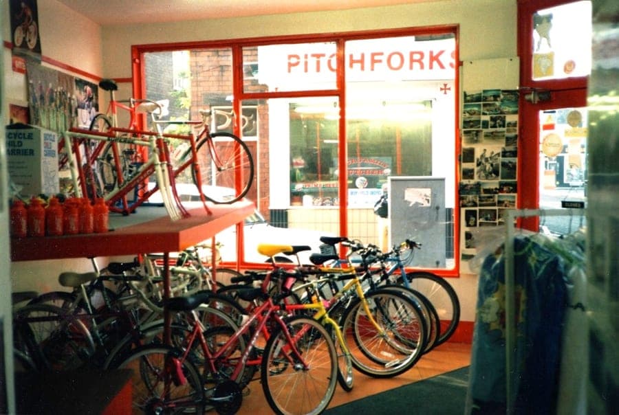 Looking out of the shop window onto the street past a row of bicycles parked inside. A shopfront across the road reads 'Pitchforks'.