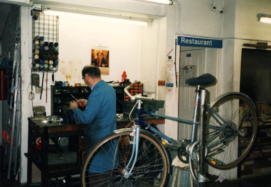 Sid at the workbench with a bicycle beside him and a doorway marked 'Restaurant' behind.