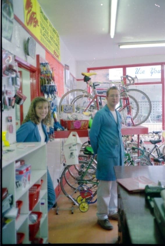 A woman and a man in a blue workshop coat standing among bikes and racked accessories inside the shop.