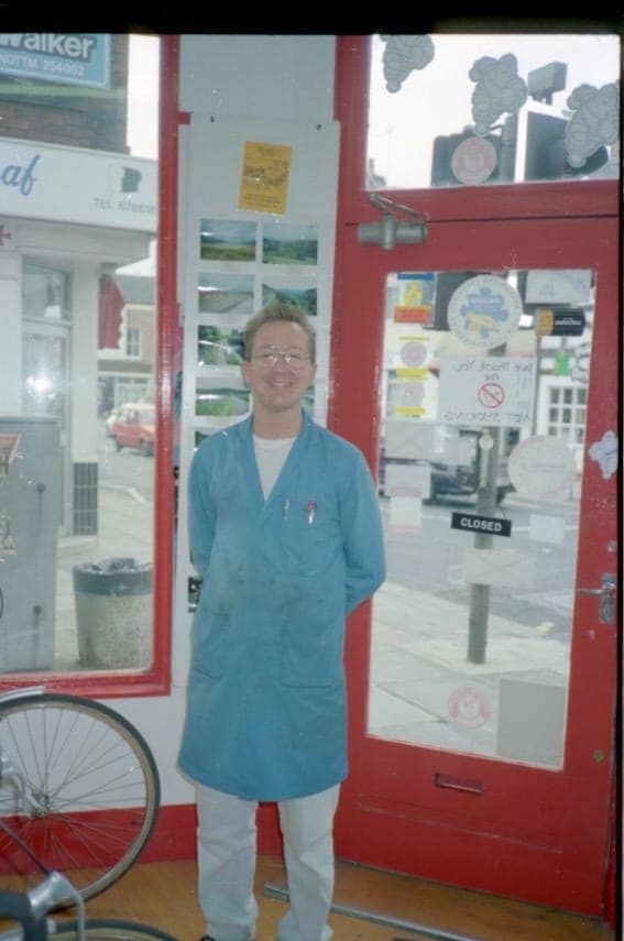 A man in a blue workshop coat standing in the open doorway of the shop, a red frame beside him and the street reflected in the glass.