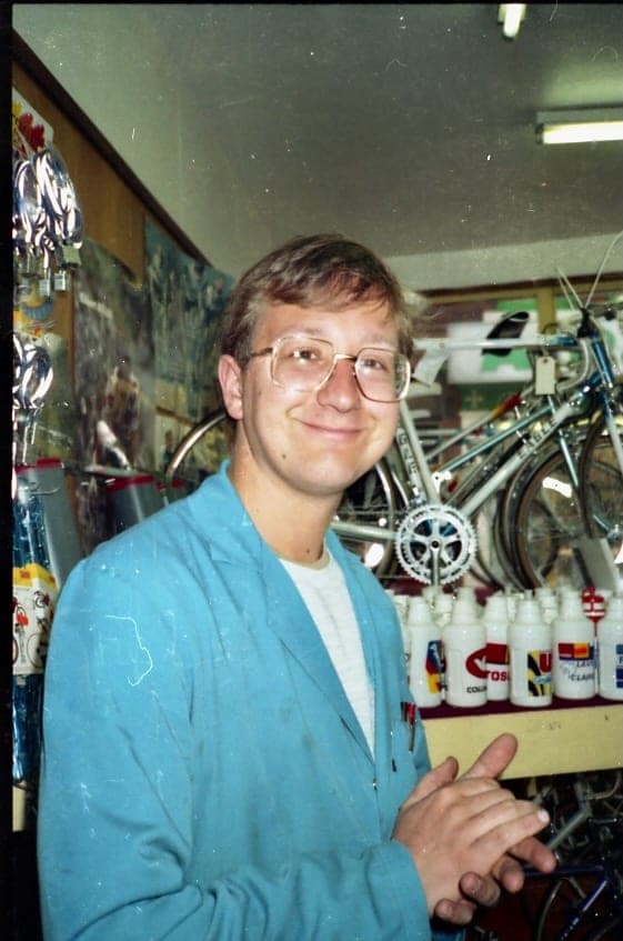 A young man in a blue shop coat standing beside a rack of bicycles, a shelf of bottles at his elbow and a cabinet of trophies on the wall behind him.