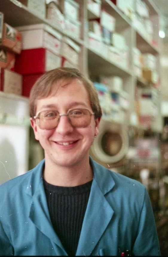 A young man in glasses and a blue shop coat smiling at the camera, the shelves of stock behind him out of focus.