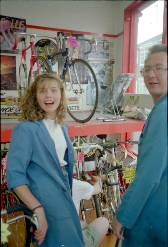 A woman and a man in a blue workshop coat mid-laugh in front of a rack of bikes and a wheel hung on the wall.