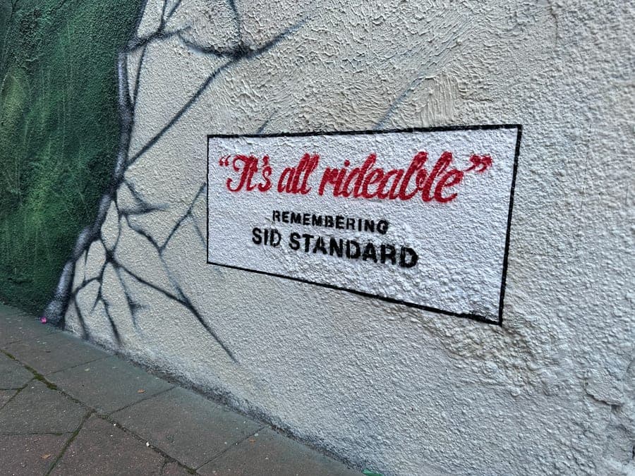 A panel painted at the foot of the mural on a rendered wall: 'It's all rideable' in red script inside a black box, with REMEMBERING SID STANDARD beneath it in black capitals.