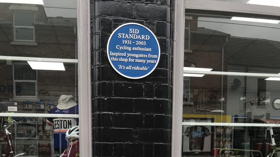 A blue circular plaque fixed to the dark brick pillar between two shop windows, bicycles and stock visible through the glass on either side.