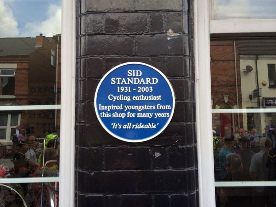 Close view of the blue plaque reading 'Sid Standard 1931-2003, cycling enthusiast, inspired youngsters from this shop for many years — It's all rideable'. A crowd of people is reflected in the shop window beside it.