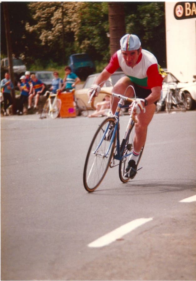 A rider in a red, white and green jersey out of the saddle on a road, spectators and parked bikes at the roadside behind.