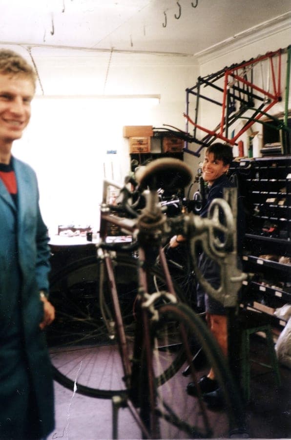 Two men in the workshop, one in a blue coat, with a bicycle held up in a repair stand between them and rims racked on the wall behind.