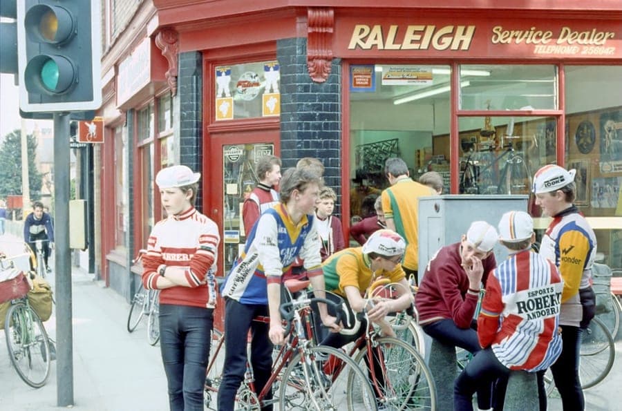 A group of young riders in club jerseys standing with their bikes on the pavement outside the shop, under a Raleigh Service Dealer sign.