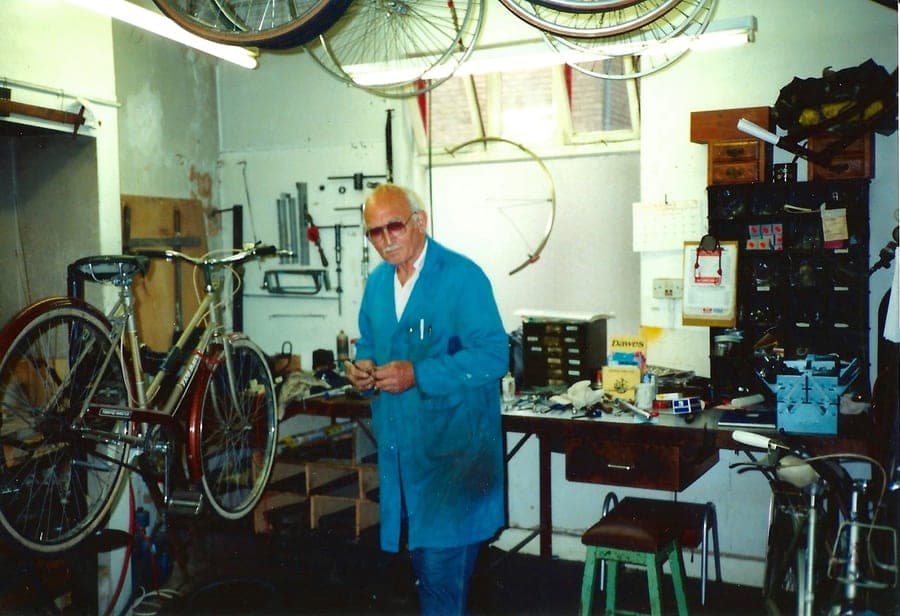 A man in a pale blue coat working at a bench in the back workshop, a finished bicycle beside him and tools racked on the wall.