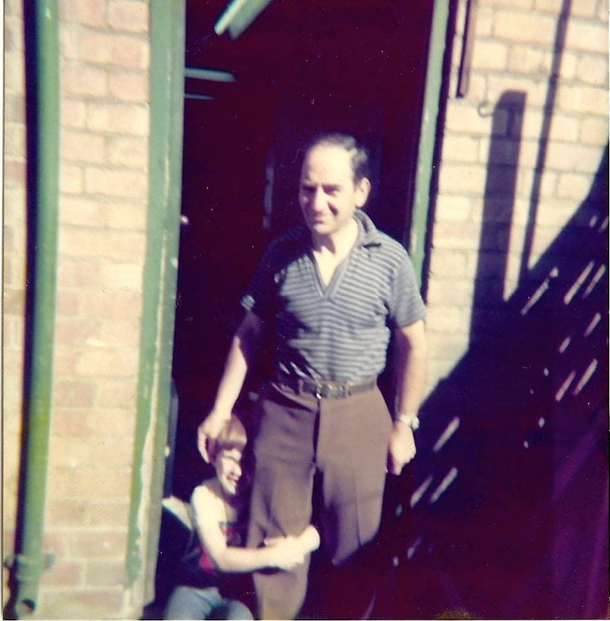 A faded colour snapshot. Sid stands in a brick doorway in a striped polo shirt, a young boy beside him.