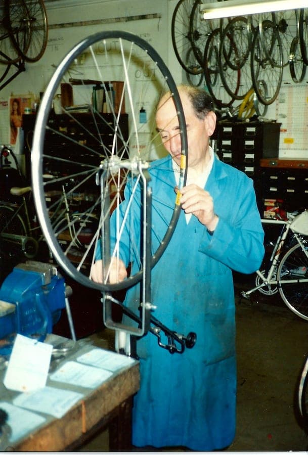 Sid in a blue coat with a wheel in a truing stand and a spoke key in his hand, wheels and rims stacked behind him.
