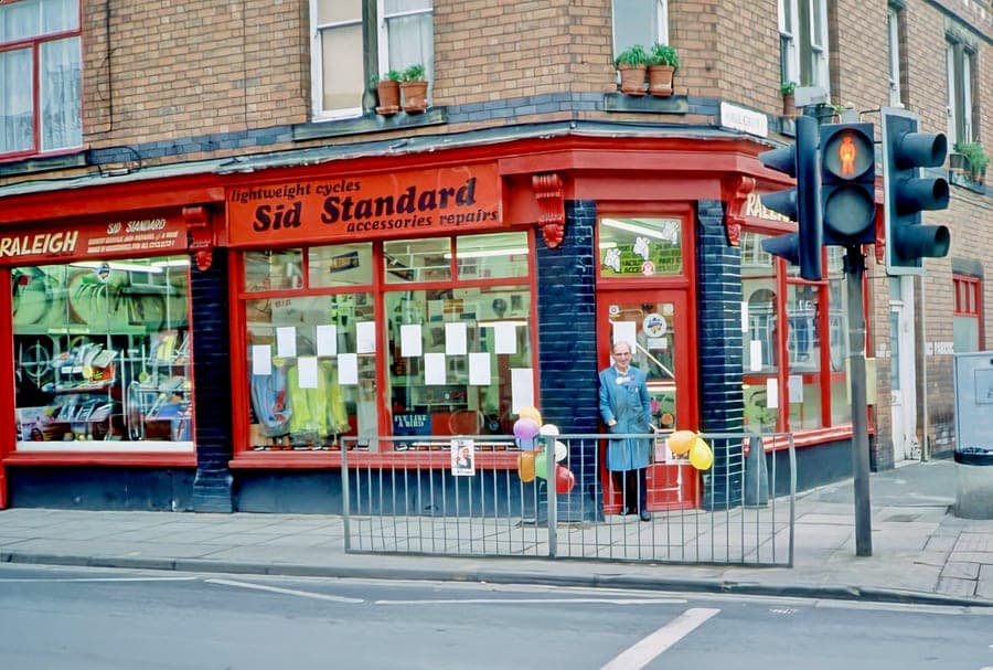 The red corner shop signwritten 'lightweight cycles — Sid Standard — accessories repairs', seen across the road junction. Someone stands in the open doorway and balloons are tied along the pavement railings.