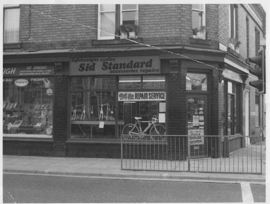 A corner shop photographed in black and white, the fascia signwritten 'lightweight cycles — Sid Standard — accessories repairs'. A bicycle stands against the railings and a Rayner repair service sign hangs in the window.