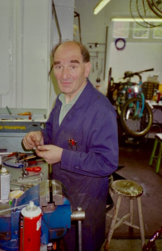 Sid in a mauve workshop coat standing at his bench, holding a small part in both hands. A wooden stool is beside him and wheels, tools and a bench light fill the wall behind.
