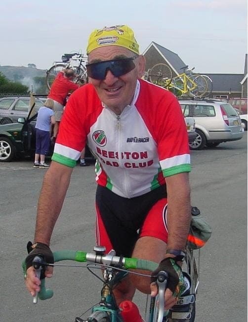 Sid on a bicycle in a red, white and green Beeston Road Club jersey, wearing sunglasses and a yellow cap, at an event with cars and other riders behind.