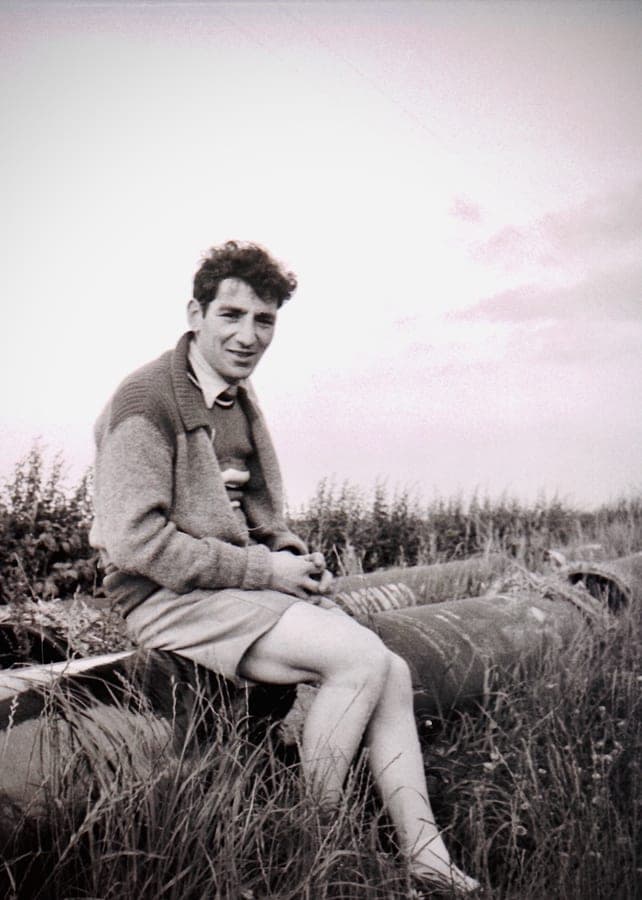 A young man in a knitted cardigan and shorts sitting in long grass, looking away from the camera, in black and white.