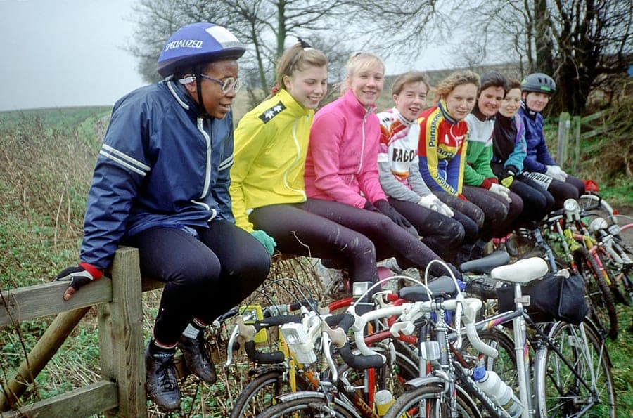 Eight teenage girls in bright cycling jackets sit in a row along a wooden fence rail on a misty winter morning, laughing and looking off to one side. Their bicycles are propped in front of them, bars and saddles filling the foreground.