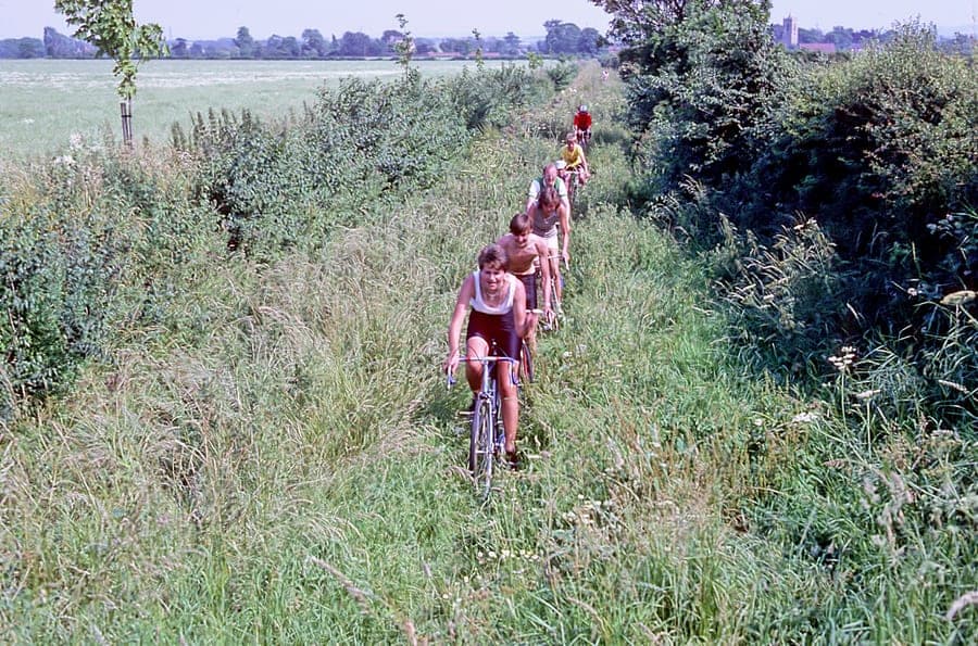 Six riders in single file coming down a narrow grassy track between waist-high summer grass and hedgerow, a church tower on the horizon behind them.