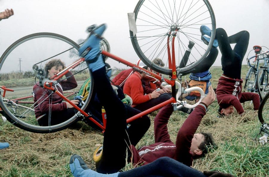 Several young riders in maroon club tracksuits lying on their backs on a grass verge with their legs in the air, holding two bicycles up above them by the wheels.