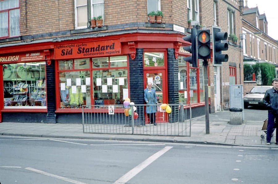 A red-painted corner shop at a road junction, signwritten 'lightweight cycles — Sid Standard — accessories repairs'. Sid stands on the pavement outside with balloons tied to the railings.