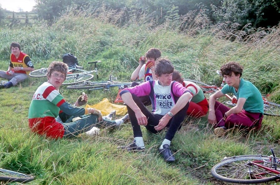 Six young riders sitting in long grass at the side of a lane, eating, with their bicycles laid down around them. One wears a green and red club jersey, another a Miko-Mercier team top.
