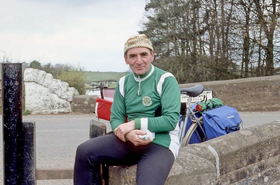 Sid sitting on a stone bridge parapet in a green and white cycling jersey and a knitted hat, holding a sandwich, with his loaded touring bicycle and blue panniers beside him.