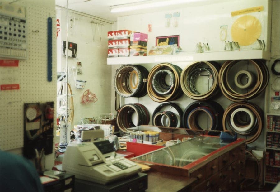 A shop counter with a cash register on it and a glass display case in front. Tubular tyres hang in loops on the wall behind, with rims stacked above them.