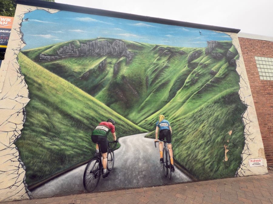 A mural painted on a gable wall so that the wall appears broken open onto a steep green Derbyshire dale with limestone crags along the top. Two cyclists in Beeston Road Club jerseys ride away up the road into it. A small painted panel at the lower right reads 'It's all rideable — remembering Sid Standard'.