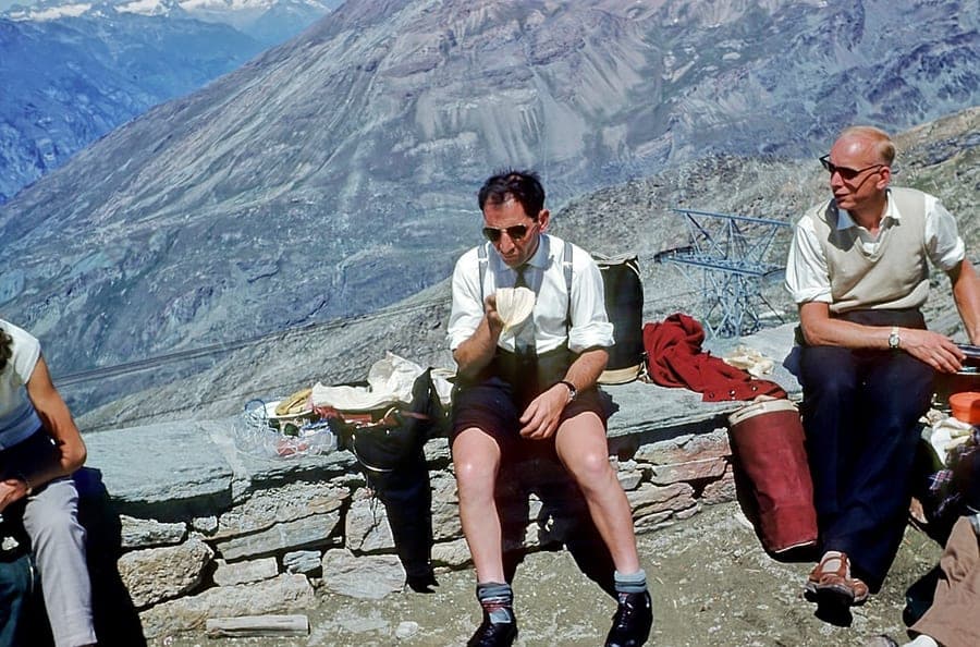 Three men sitting on a low stone wall eating, with a bare scree mountainside rising behind them and panniers on the ground.