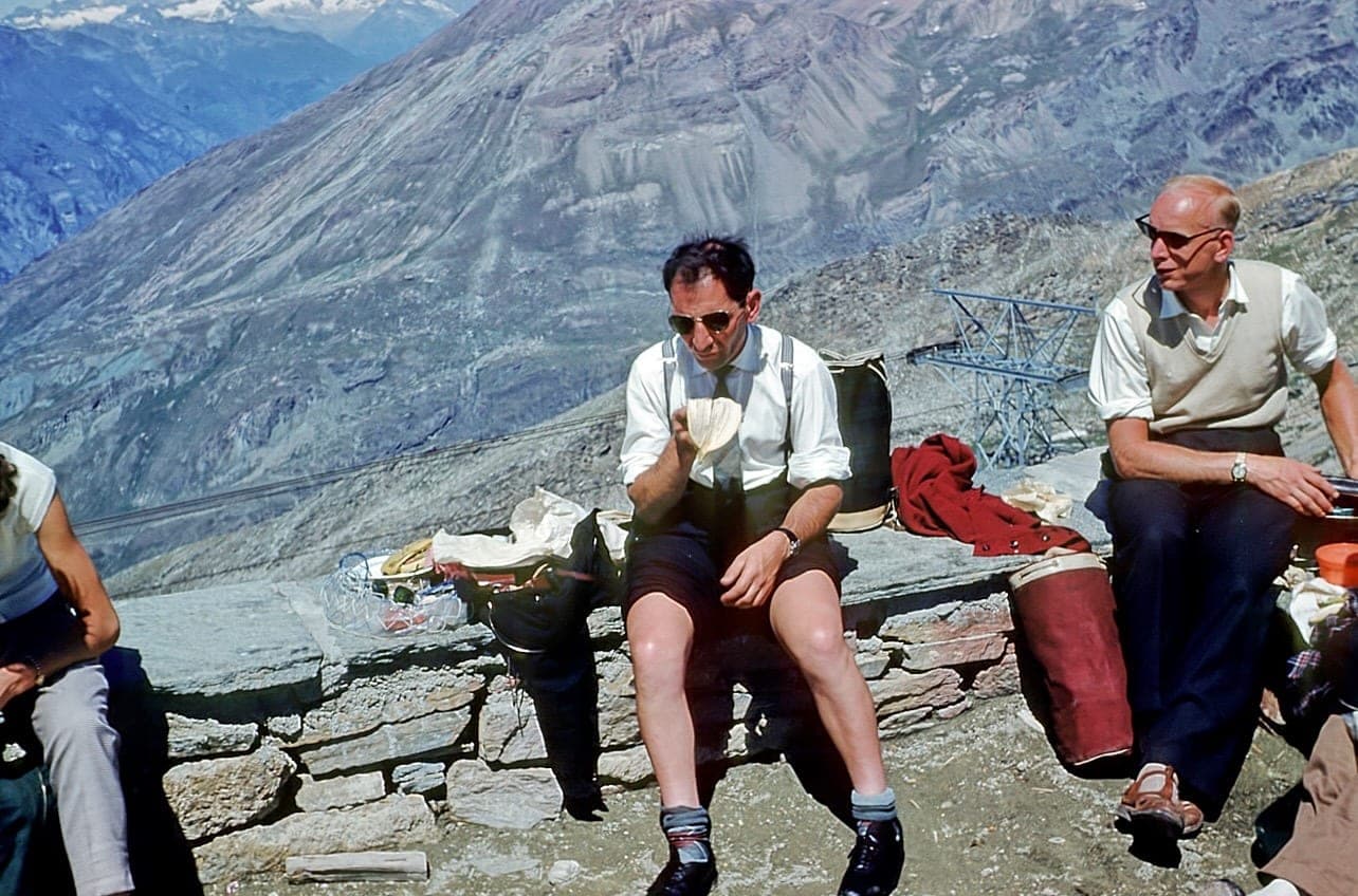 Three men sitting on a low stone wall eating, with a bare scree mountainside rising behind them and panniers on the ground.