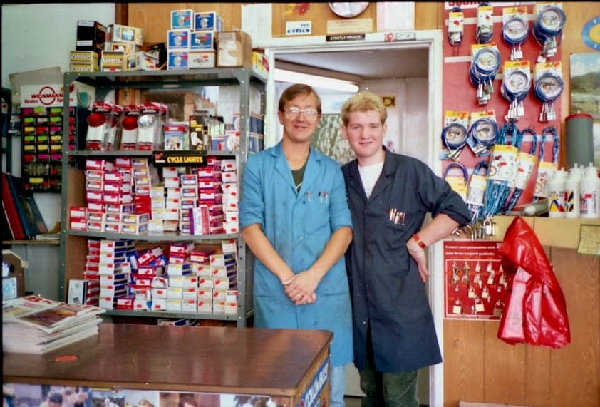 Two young men in shop coats, one blue and one dark, standing together in a doorway between shelves stacked with boxed accessories, pumps and lights.