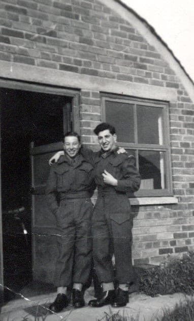 Two young men in battledress standing arm in arm against a brick wall beside a doorway, in black and white.