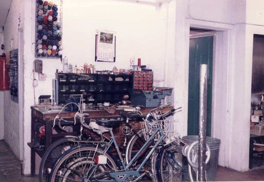 A back workshop with a cluttered bench along the wall, jars and tins on shelves above it, and part-built bicycles standing on the floor.